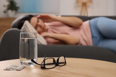 Sad woman suffering from headache indoors, focus on glass of water, pills and eyeglasses