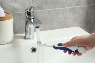 Woman washing electric toothbrush under flowing water from faucet in bathroom, closeup