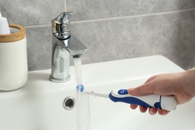 Photo of Woman washing electric toothbrush under flowing water from faucet in bathroom, closeup