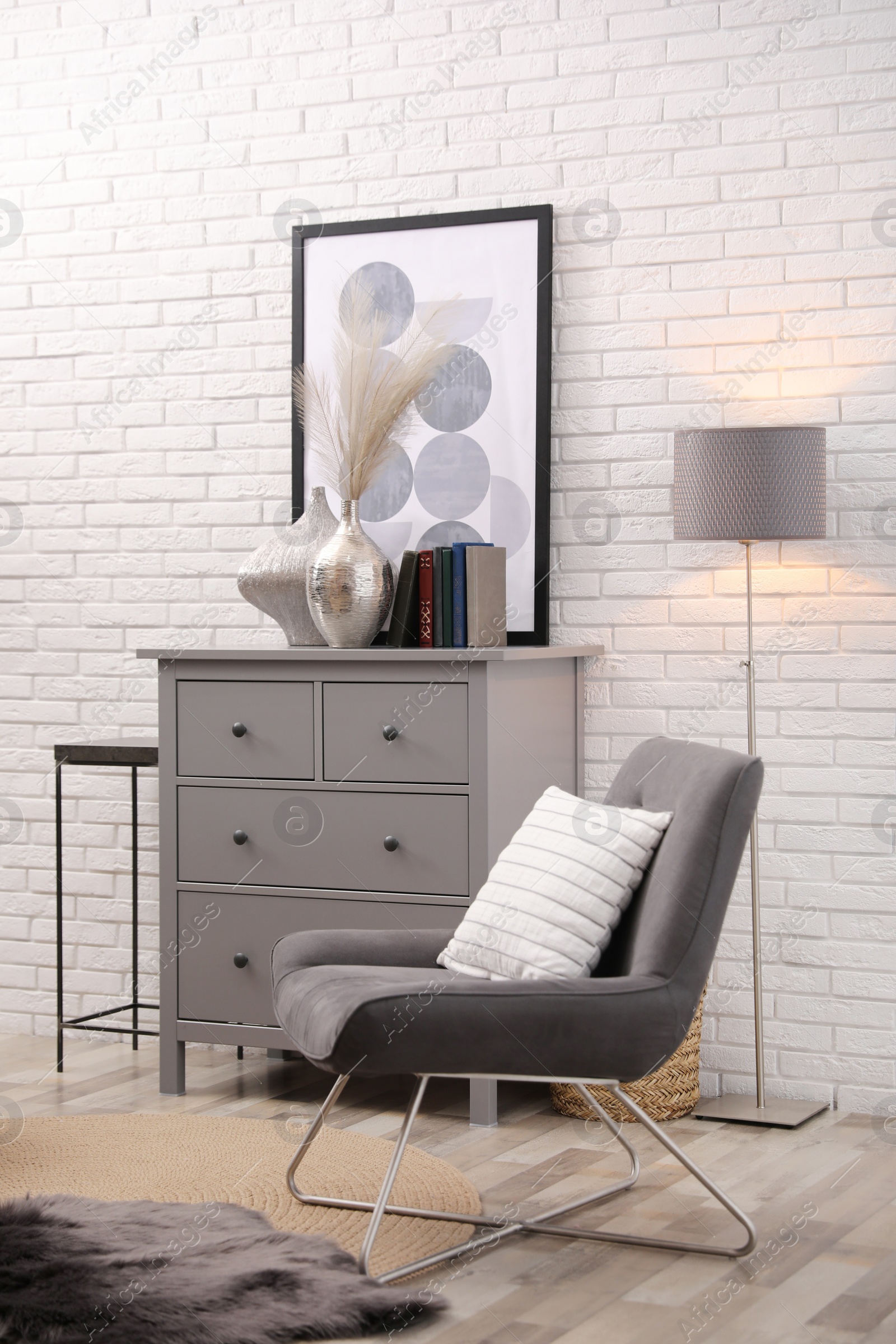 Photo of Stylish room interior with grey chest of drawers and chair near white brick wall