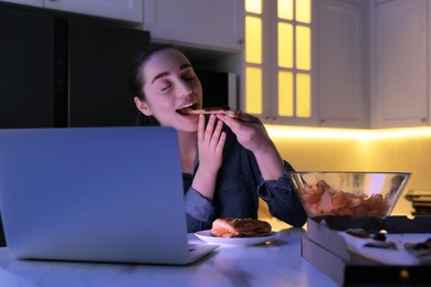 Young woman eating pizza while using laptop in kitchen at night. Bad habit