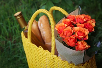 Yellow wicker bag with beautiful roses, bottle of wine and baguettes on green grass outdoors, closeup