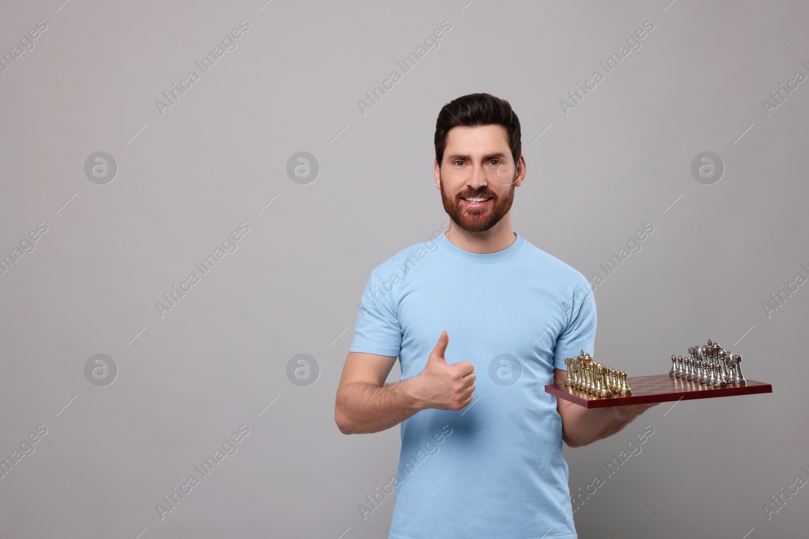 Photo of Smiling man holding chessboard with game pieces and showing thumb up on light grey background. Space for text