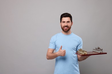 Smiling man holding chessboard with game pieces and showing thumb up on light grey background. Space for text