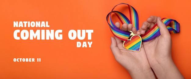 Image of National Coming Out day, October 11. Woman holding rainbow ribbon with heart shaped pendant on orange background, top view. LGBT pride