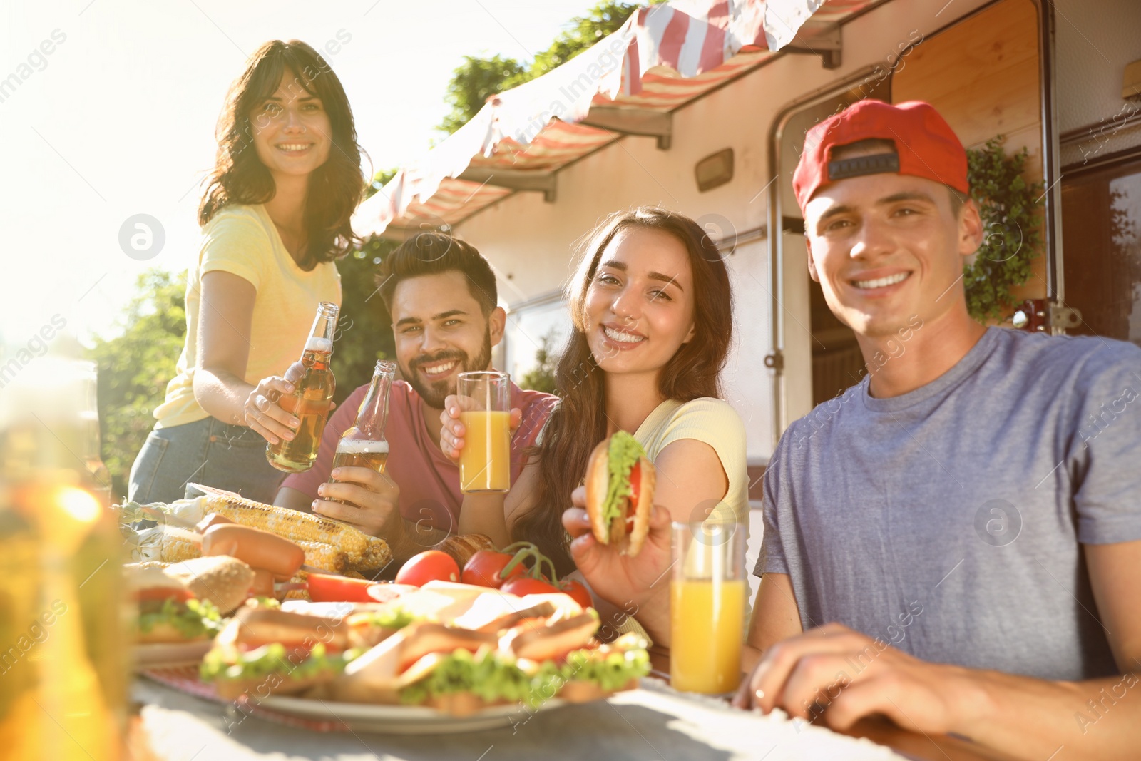 Photo of Happy friends with drinks and food at table near motorhome. Camping season