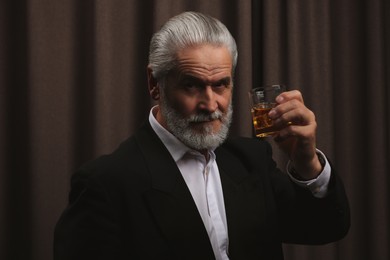 Senior man in formal suit holding glass of whiskey with ice cubes on brown background