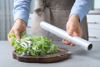 Photo of Woman putting plastic food wrap over fresh lettuce at light grey table indoors, closeup