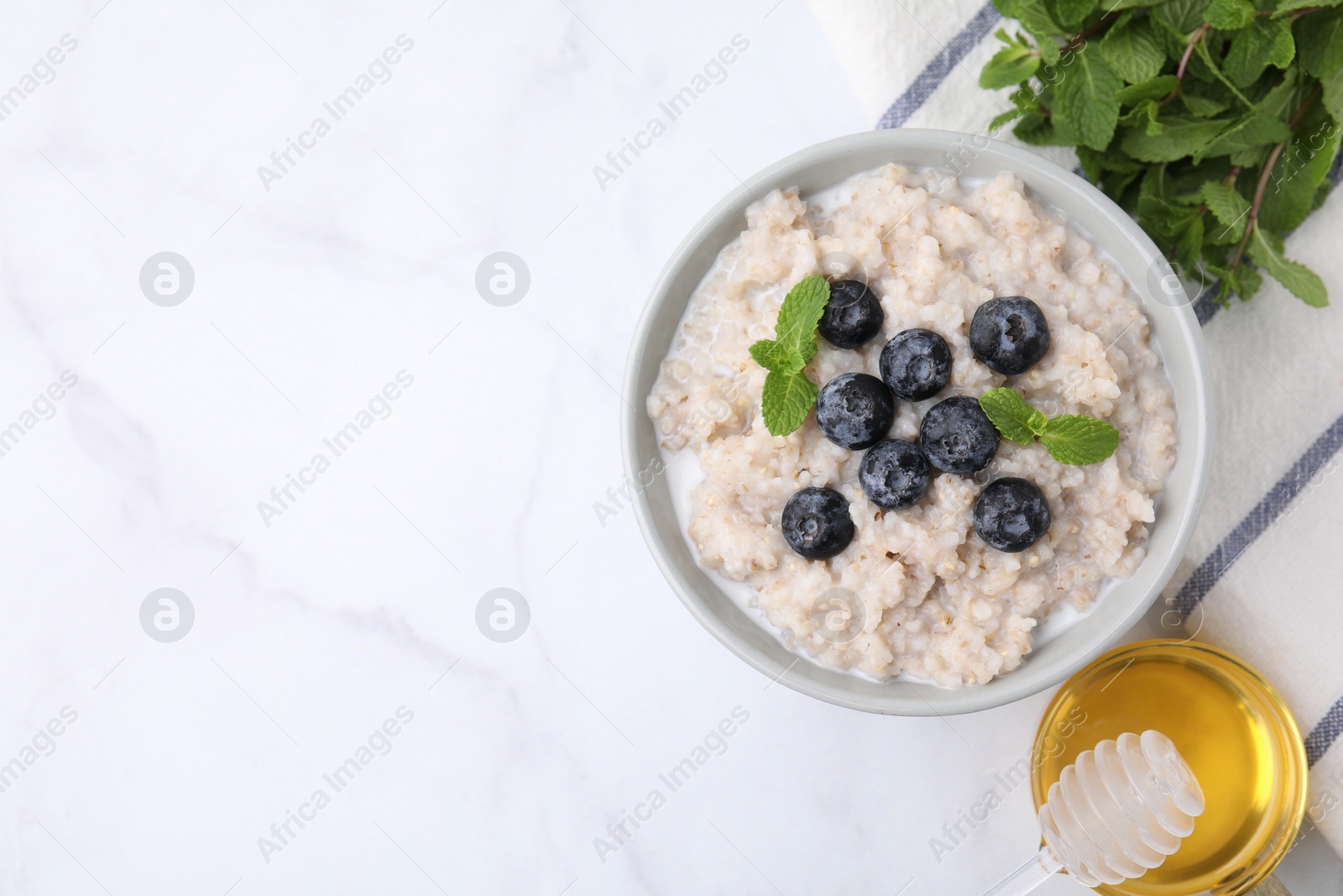 Photo of Delicious barley porridge with blueberries and mint in bowl served with honey on white marble table, flat lay. Space for text