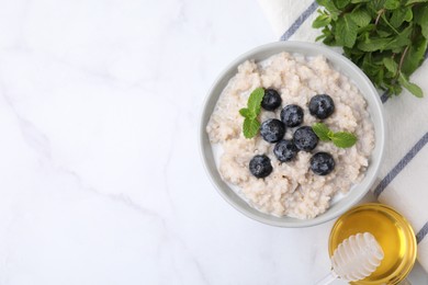 Delicious barley porridge with blueberries and mint in bowl served with honey on white marble table, flat lay. Space for text