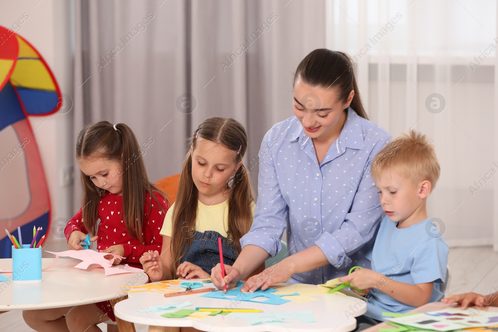 Photo of Nursery teacher and group of cute little children making toys from color paper at desks in kindergarten. Playtime activities