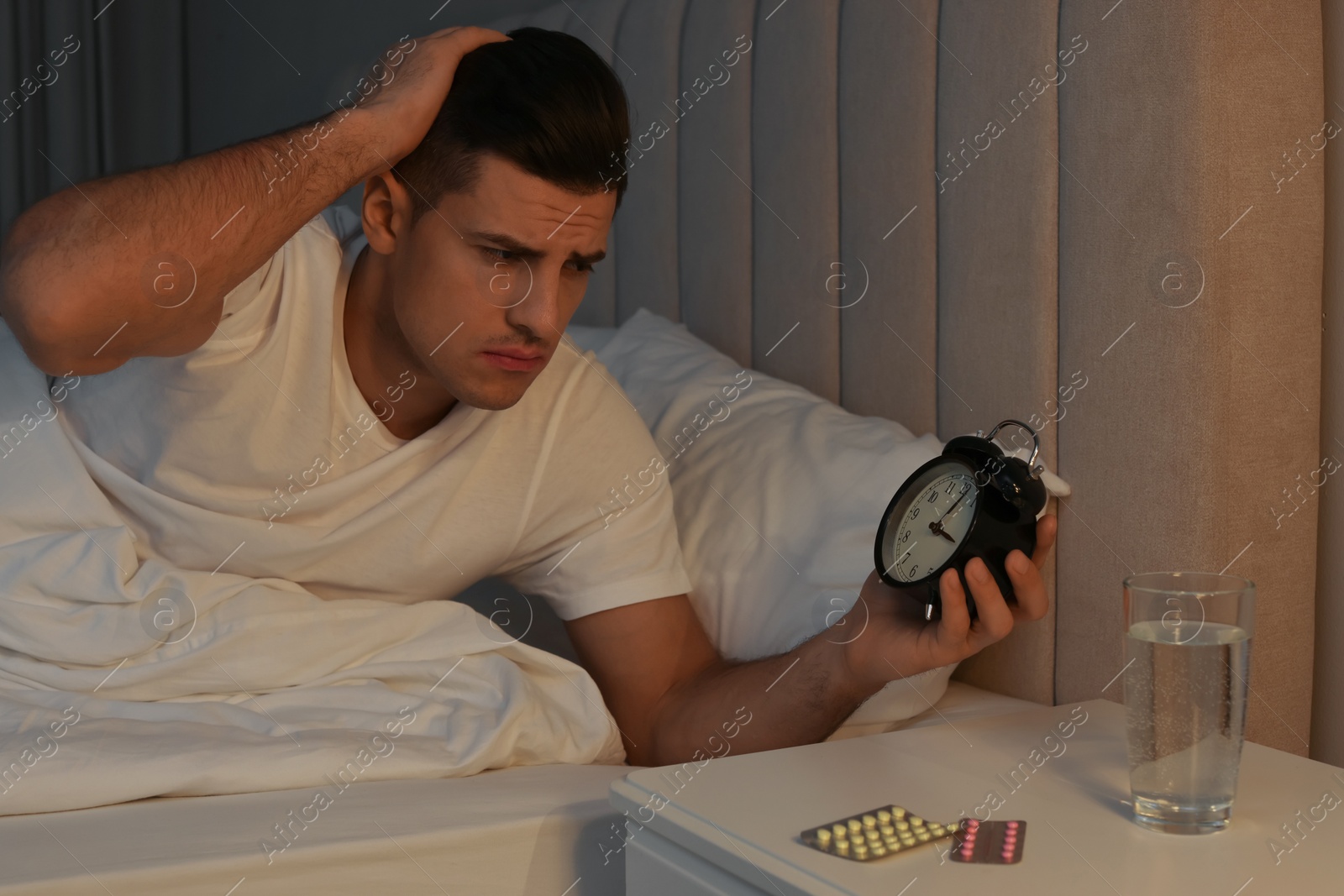 Photo of Man suffering from insomnia looking at time on alarm clock in bed indoors