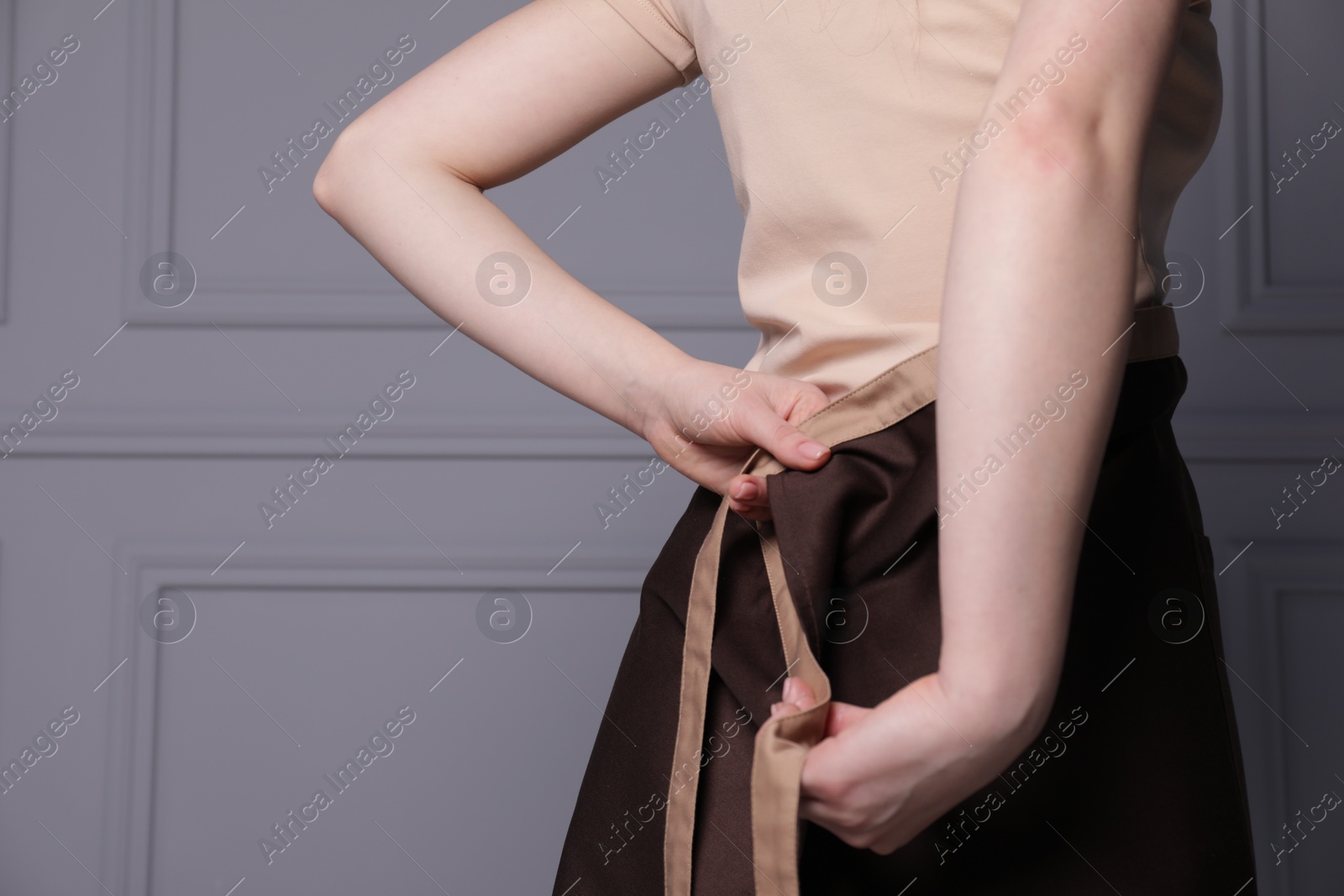 Photo of Woman putting on brown apron against grey wall, closeup. Space for text