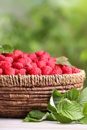 Photo of Wicker basket with tasty ripe raspberries and leaves on white wooden table against blurred green background, closeup. Space for text