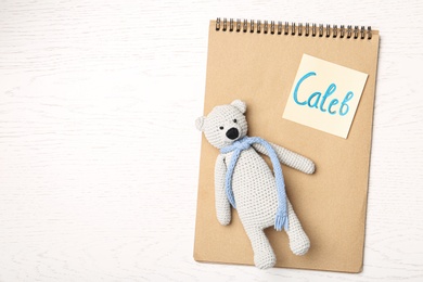 Photo of Notebook with written baby name and toy on white wooden table, top view. Space for text