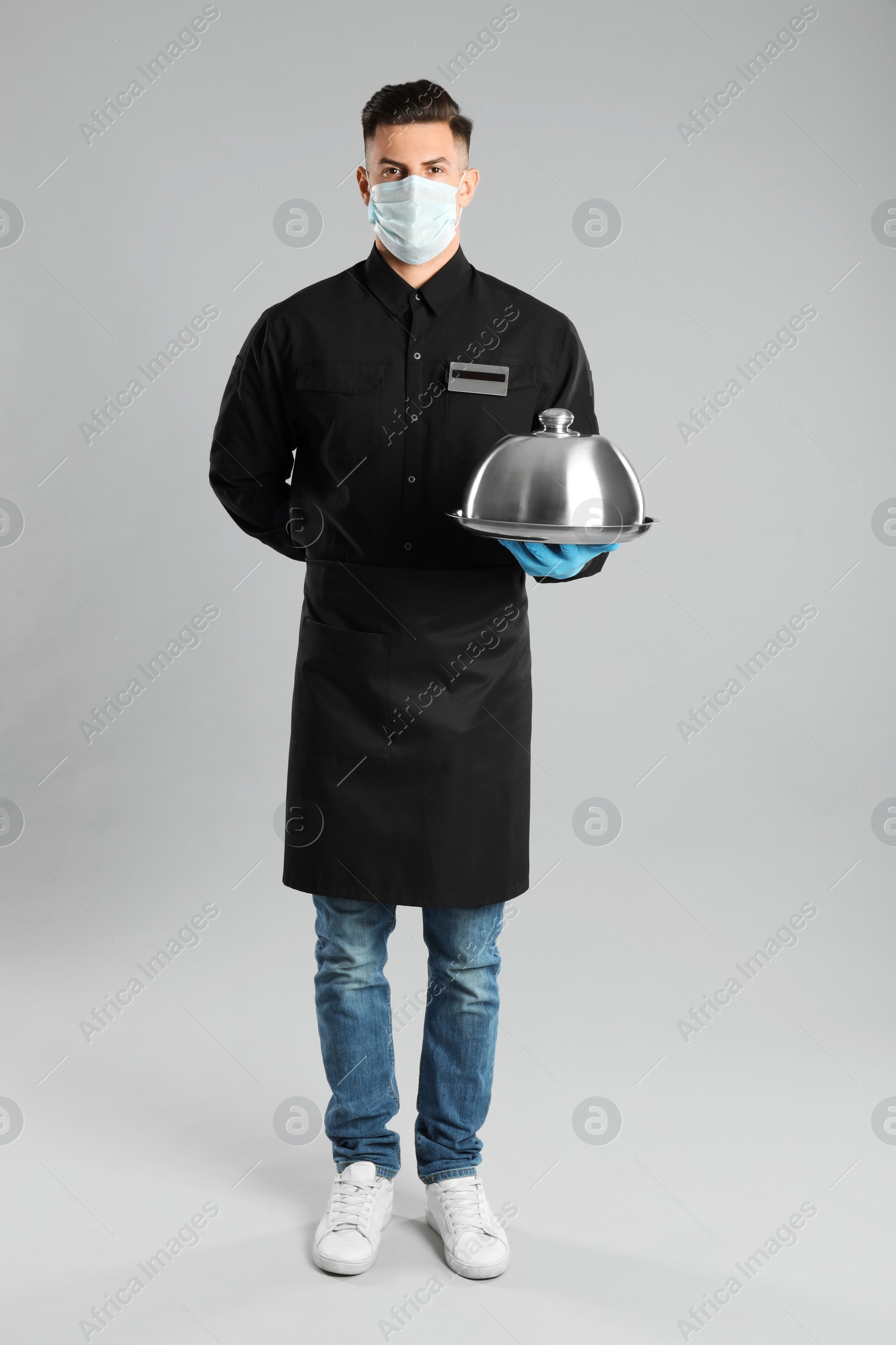 Photo of Waiter in medical face mask holding tray with lid on light grey background