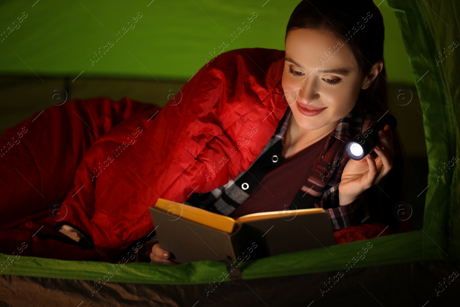 Photo of Young woman with flashlight reading book in tent