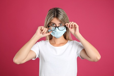 Photo of Woman wiping foggy glasses caused by wearing disposable mask on pink background. Protective measure during coronavirus pandemic