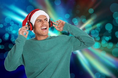 Happy man in Santa hat listening to Christmas music with headphones on bright background, bokeh effect