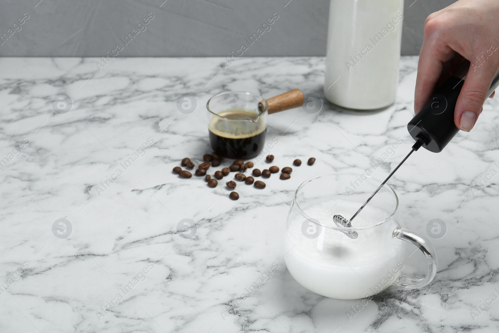 Photo of Woman whisking milk in cup with mini mixer (milk frother) at white marble table, closeup. Space for text