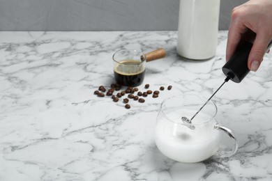 Photo of Woman whisking milk in cup with mini mixer (milk frother) at white marble table, closeup. Space for text