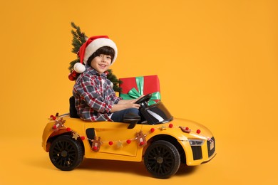 Photo of Cute little boy in Santa hat with Christmas tree and gift box driving children's electric toy car on yellow background