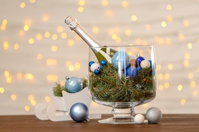 Photo of Bottle of champagne, fir tree branches and decor in glass vase on table. Christmas composition against blurred lights