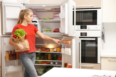 Photo of Beautiful young woman with healthy food in paper bag near refrigerator at home