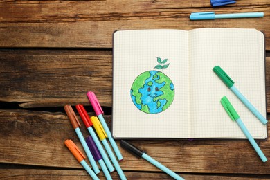 Photo of Happy Earth Day. Notebook with drawing of planet and markers on wooden table, flat lay