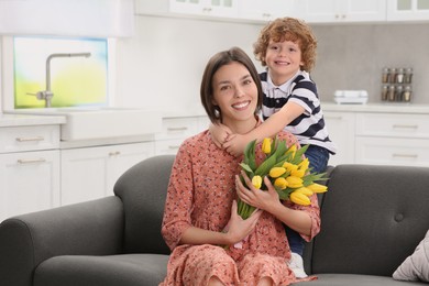 Photo of Little son congratulating his mom with Mother`s day at home, space for text. Woman holding bouquet of yellow tulips