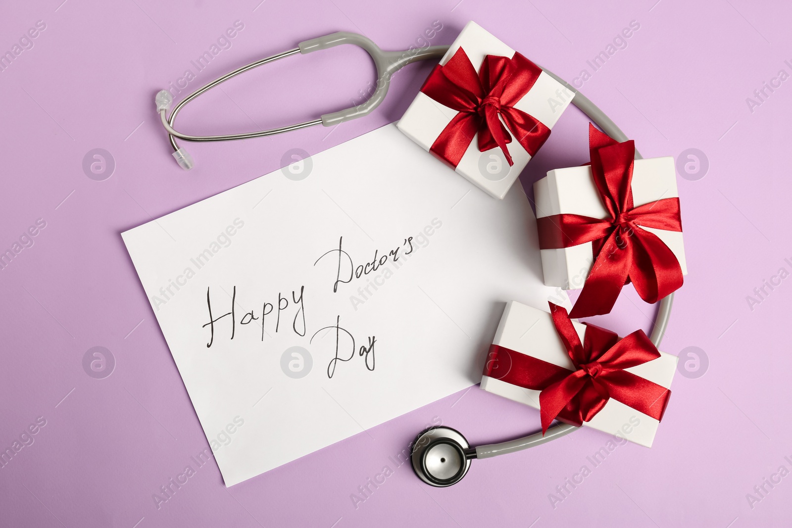 Photo of Card with phrase Happy Doctor's Day, stethoscope and gift boxes on purple background, flat lay
