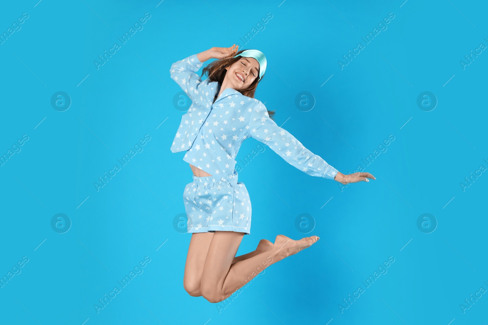 Photo of Beautiful woman with sleep mask jumping on light blue background. Bedtime