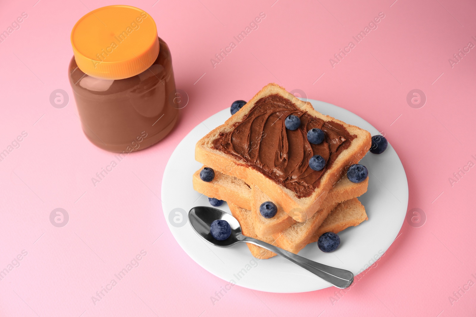 Photo of Tasty toast with chocolate paste and blueberries near jar on pink table