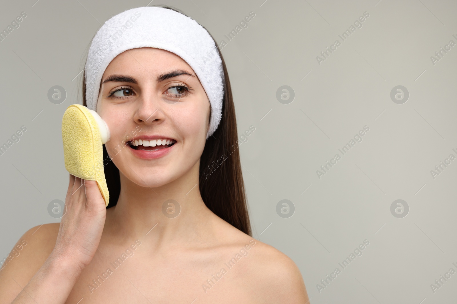 Photo of Young woman with headband washing her face using sponge on light grey background, space for text