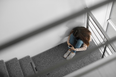 Photo of Upset teenage girl with smartphone sitting on staircase indoors, above view