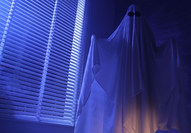 Photo of Creepy ghost. Woman covered with sheet near window in blue light, low angle view