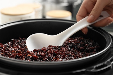 Photo of Woman taking brown rice from multi cooker with spoon in kitchen, closeup