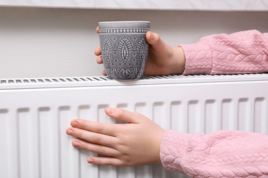 Photo of Girl with cup of drink warming hands on heating radiator indoors, closeup