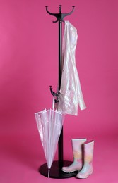 Photo of Stylish rack with umbrella, raincoat and rubber boots on pink background