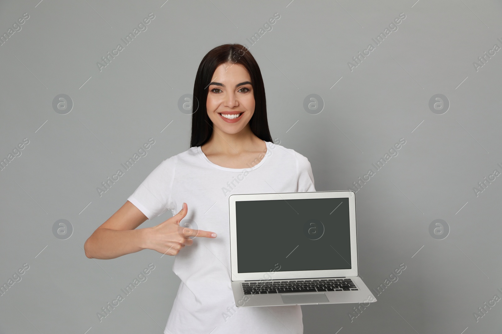 Photo of Young woman pointing at modern laptop with blank screen on light grey background