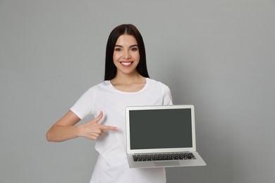 Young woman pointing at modern laptop with blank screen on light grey background