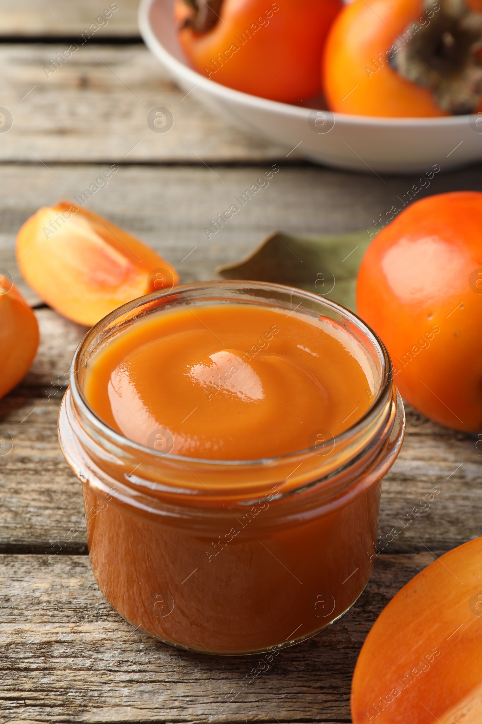 Photo of Delicious persimmon jam in glass jar and fresh fruits on wooden table, closeup