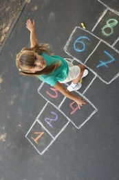 Little girl playing hopscotch drawn with chalk on asphalt outdoors, above view. Happy childhood