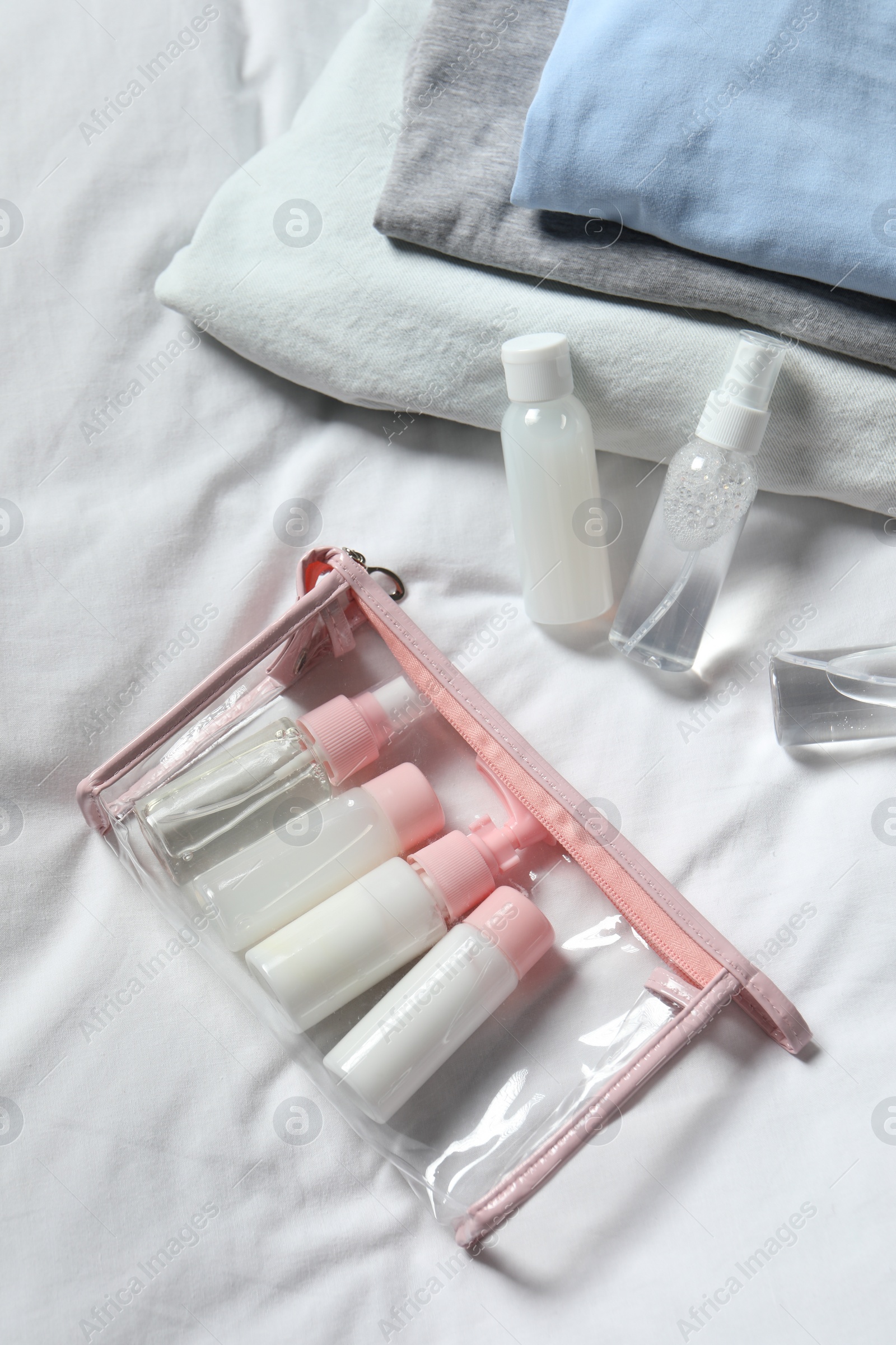 Photo of Cosmetic travel kit. Plastic bag with small containers of personal care products and stack of clothes on bed, above view