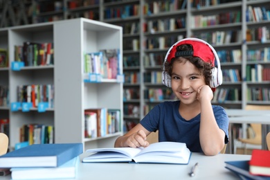 Photo of Cute little boy with headphones reading books at table in library