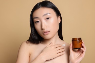 Photo of Beautiful young Asian woman with jar of body cream on beige background