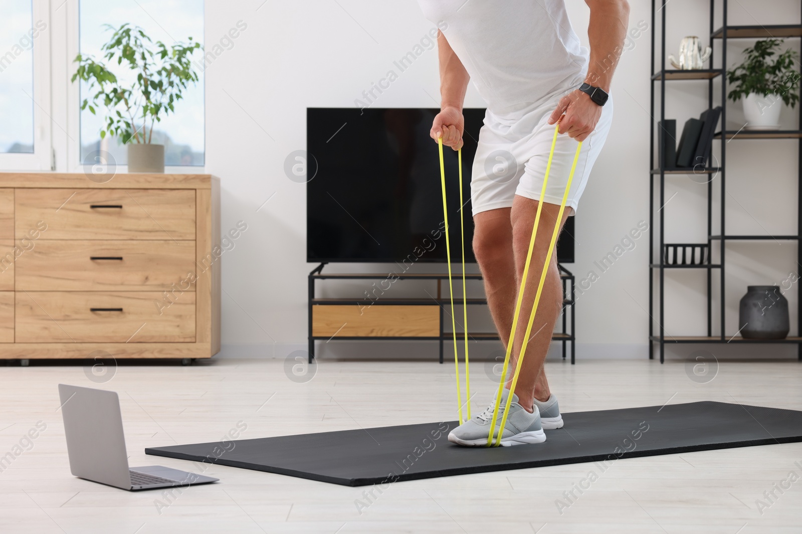 Photo of Athletic man doing exercise with elastic resistance band near laptop on mat at home, closeup