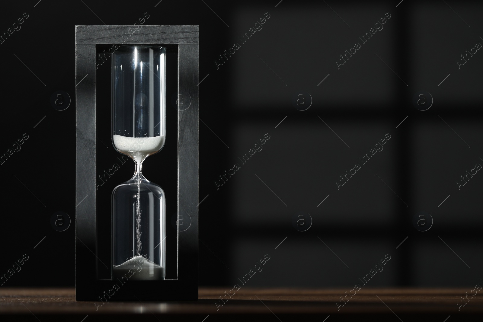 Photo of Hourglass with flowing sand on wooden table against black background, space for text