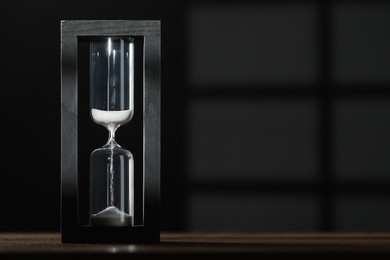 Photo of Hourglass with flowing sand on wooden table against black background, space for text