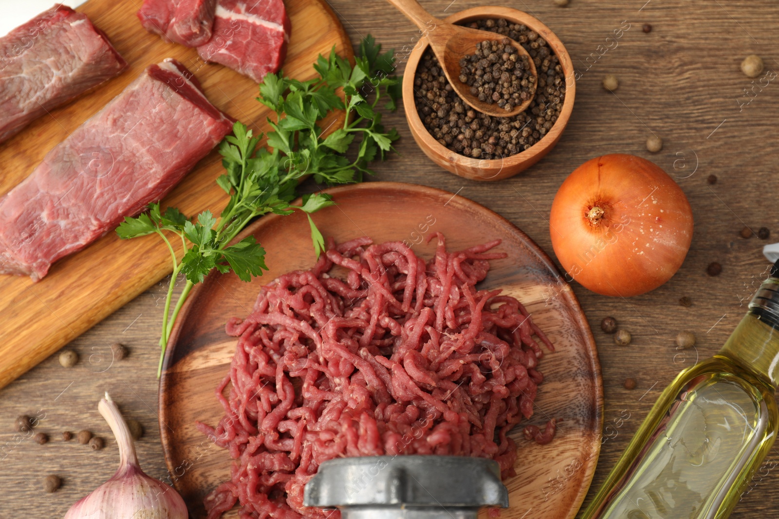 Photo of Meat grinder with beef, garlic, onion, parsley, oil and peppercorns on wooden table, flat lay